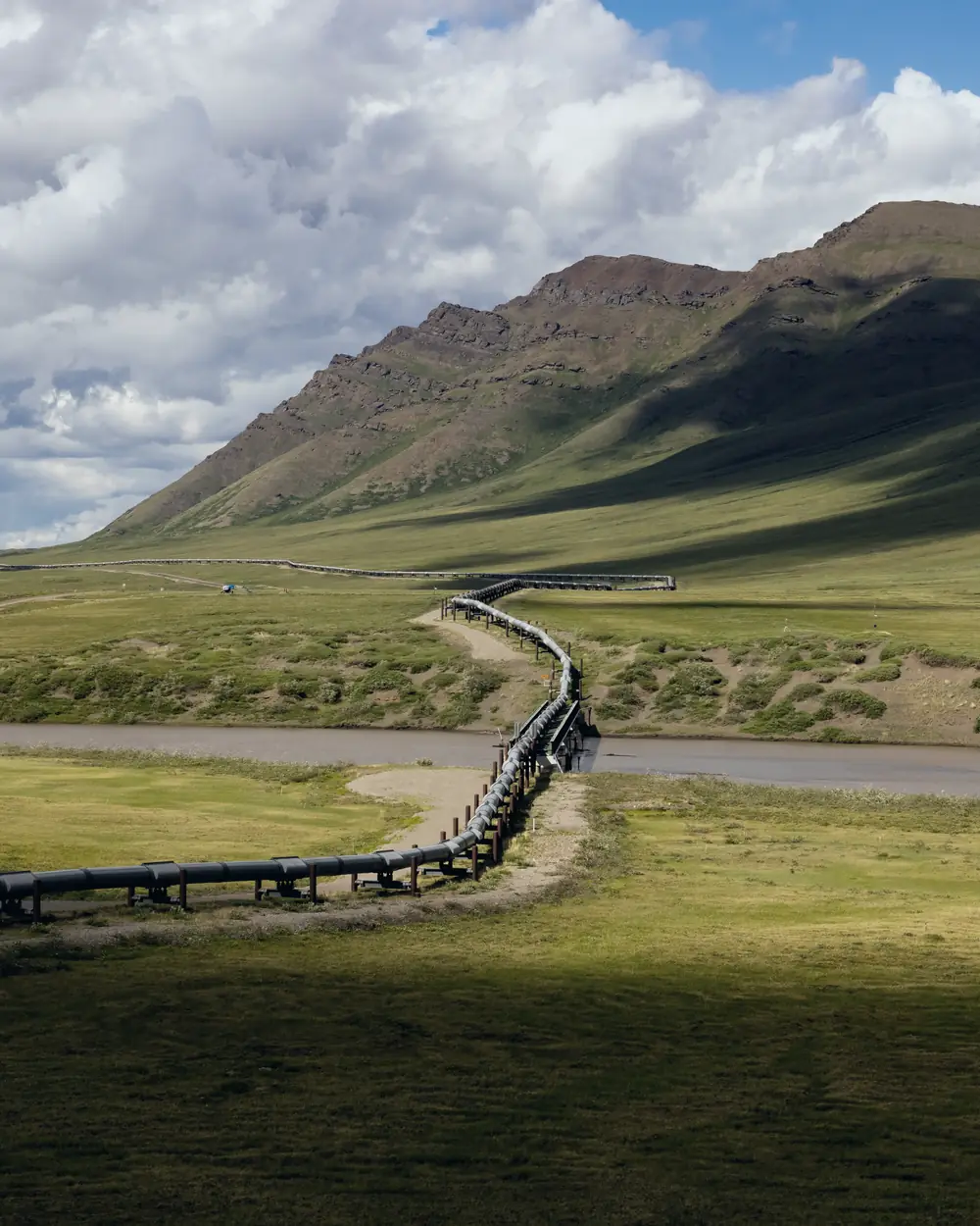 An oil pipeline curving through a green river valley below a mountain ridge